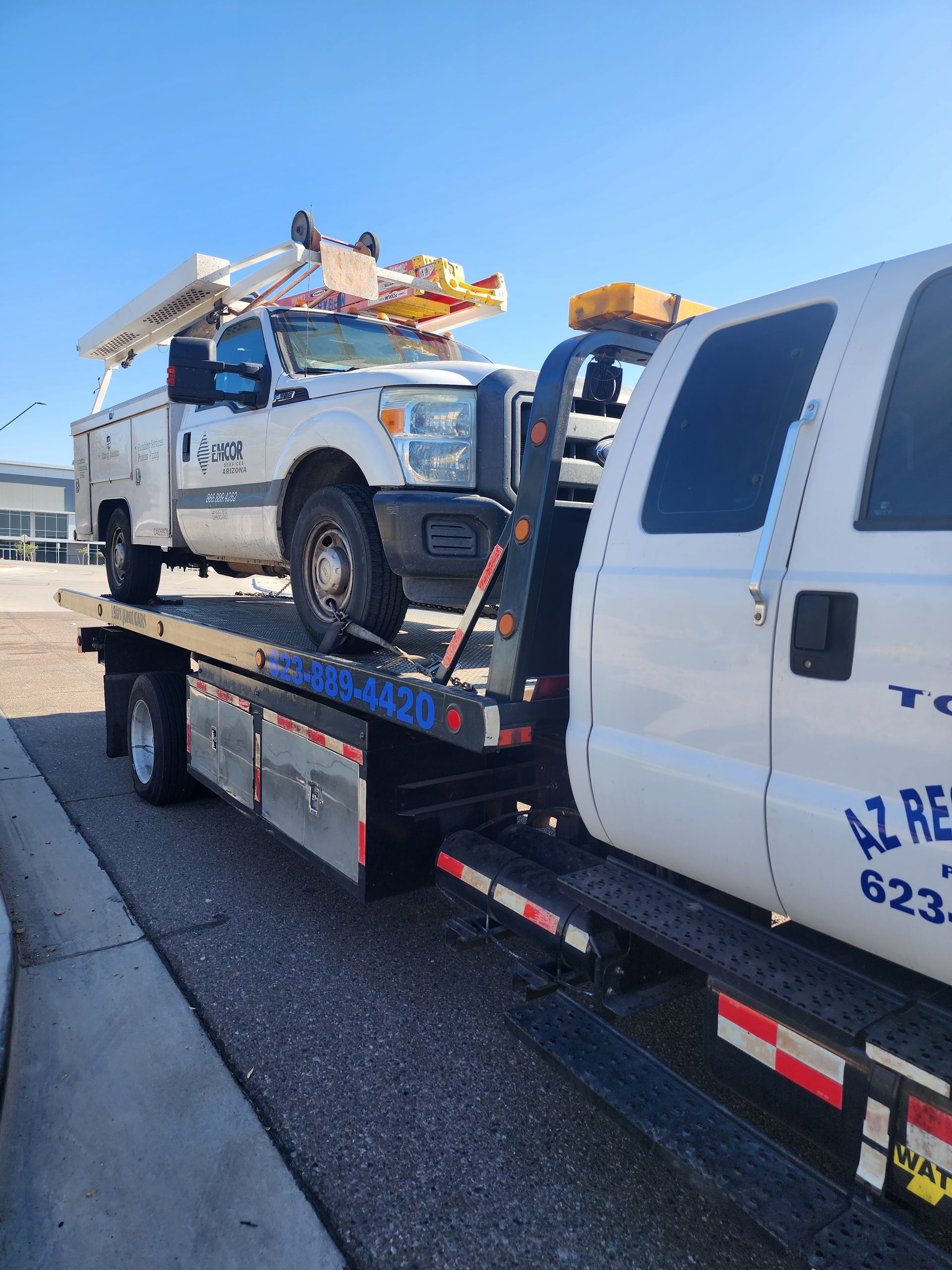 A damaged silver car being towed by a red tow truck on a residential street. Bright, sunny day.