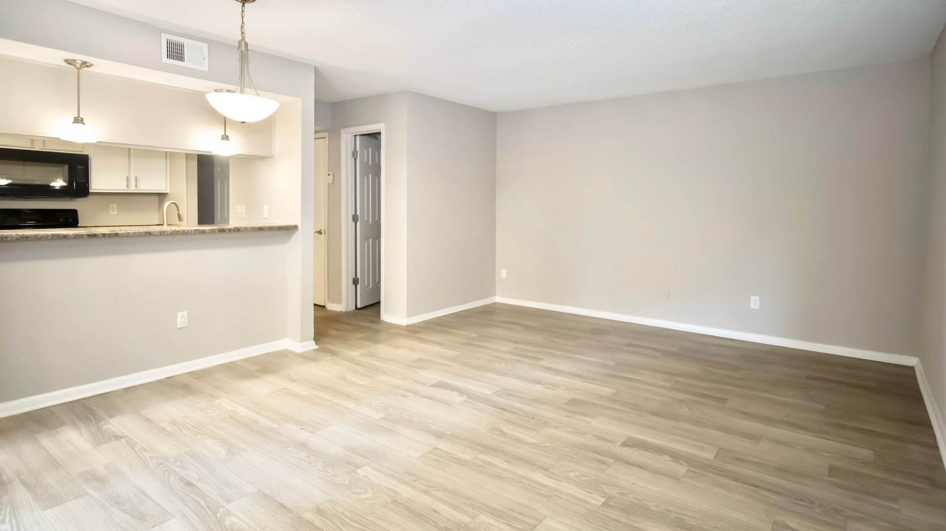 An empty living room with hardwood floors and a kitchen in the background.