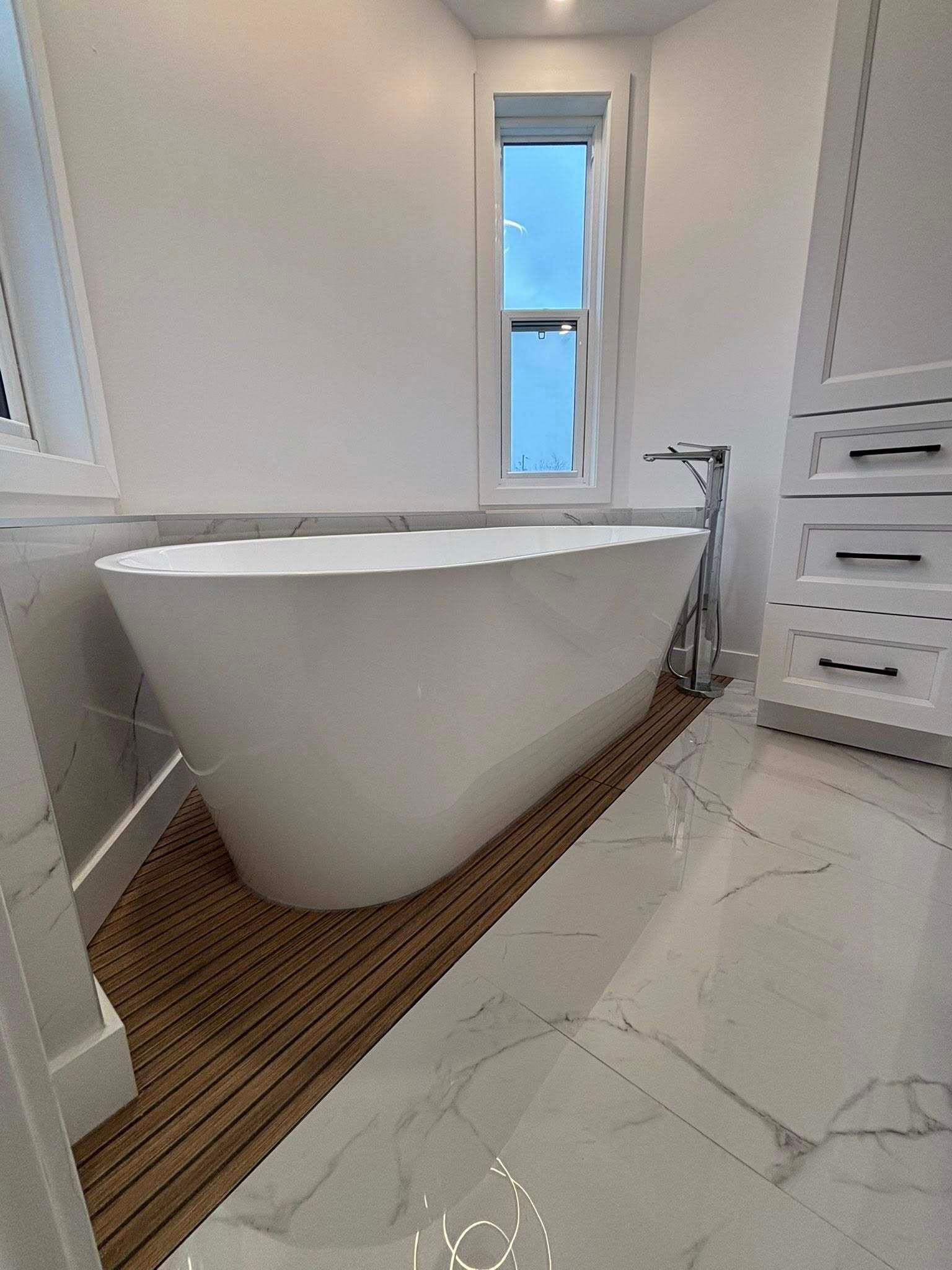 Salle de bains moderne et blanche avec baignoire îlot sur lattes de bois. Sol en marbre, meuble-lavabo et haute fenêtre.