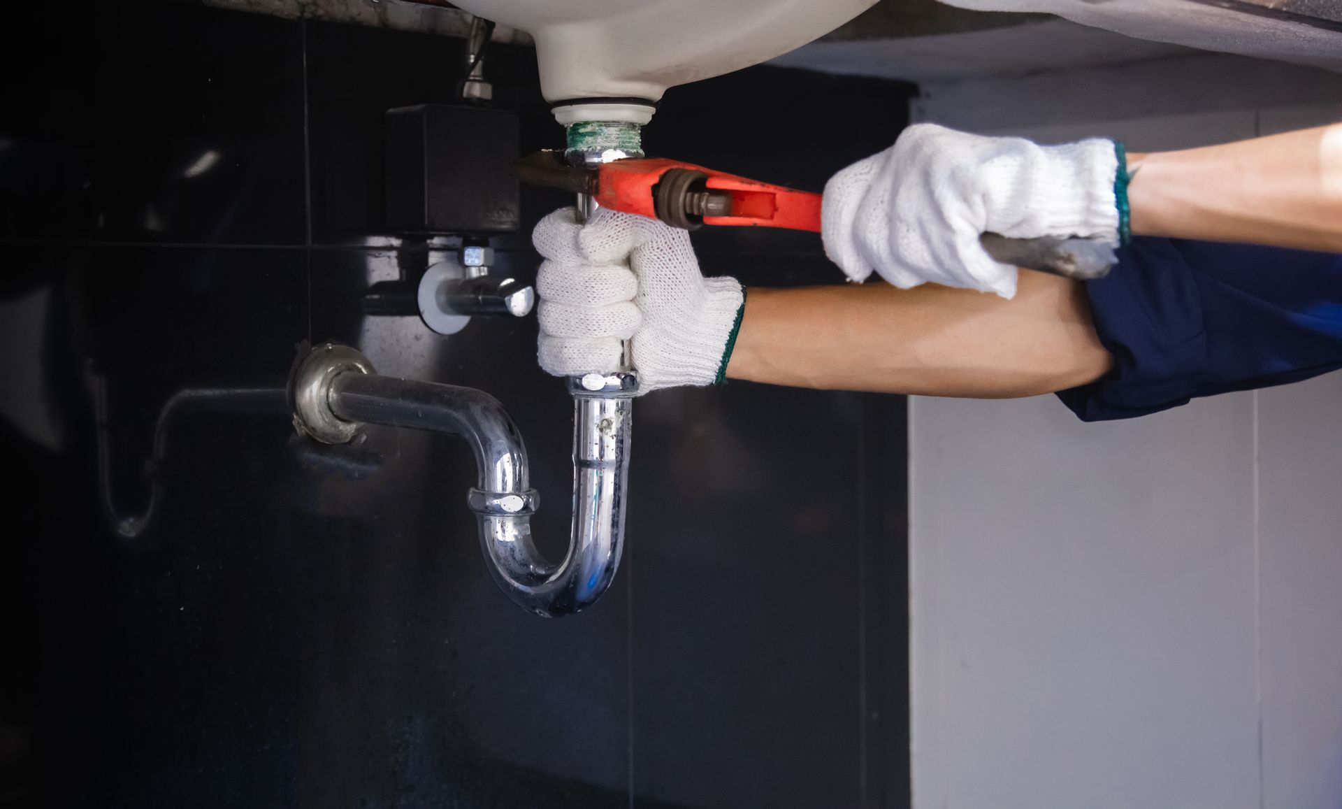 A plumber in white gloves using a wrench to adjust a sink drain pipe under a black and white sink.