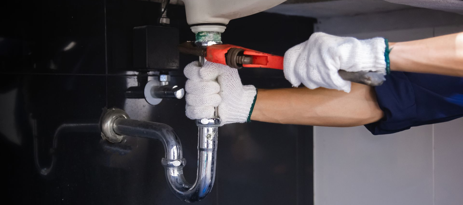 Plumber in gloves using a wrench to repair a sink's drainpipe under a black sink.