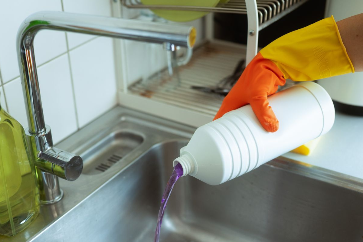 Person pouring purple liquid cleaner into a kitchen sink, wearing gloves.