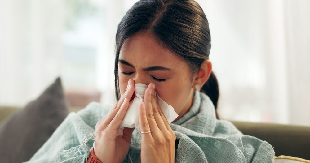 A woman is blowing her nose into a napkin while sitting on a couch.