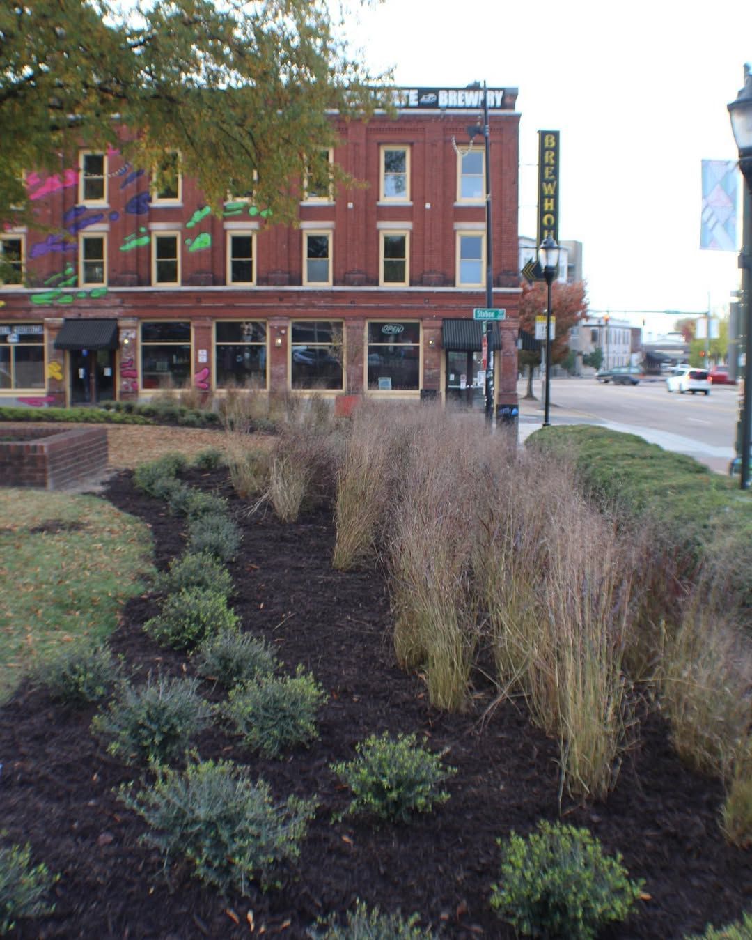 Street view with brick building, businesses, landscaping, and cars on road.