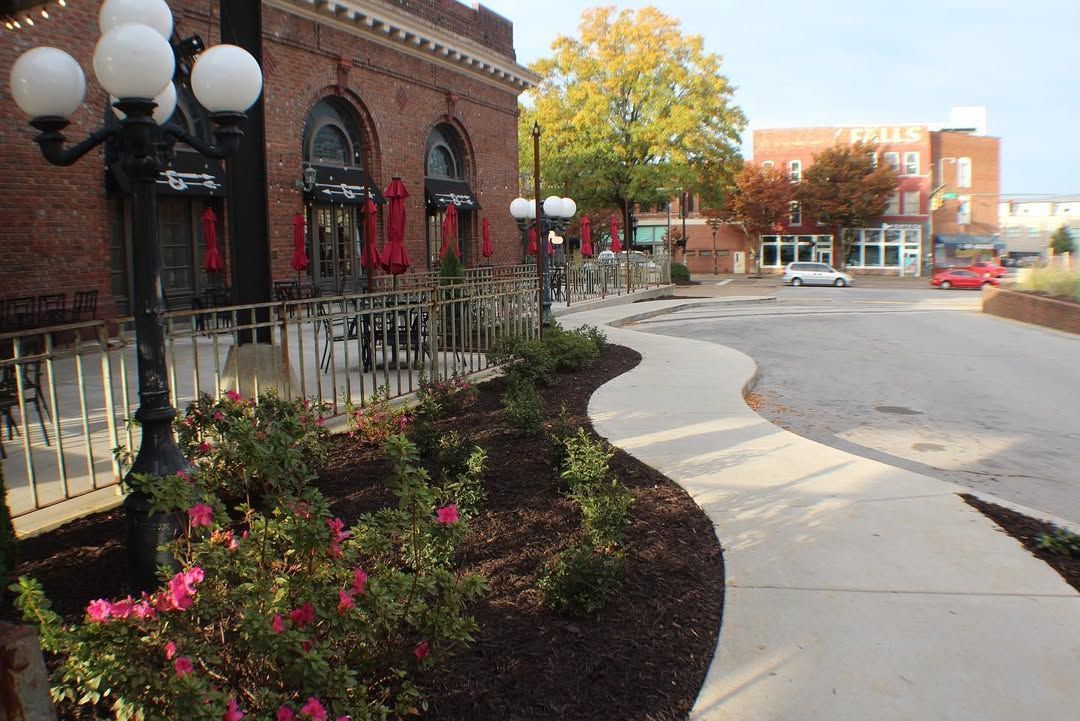 Sidewalk curves along a brick building with outdoor seating and landscaping; another building visible in the distance.