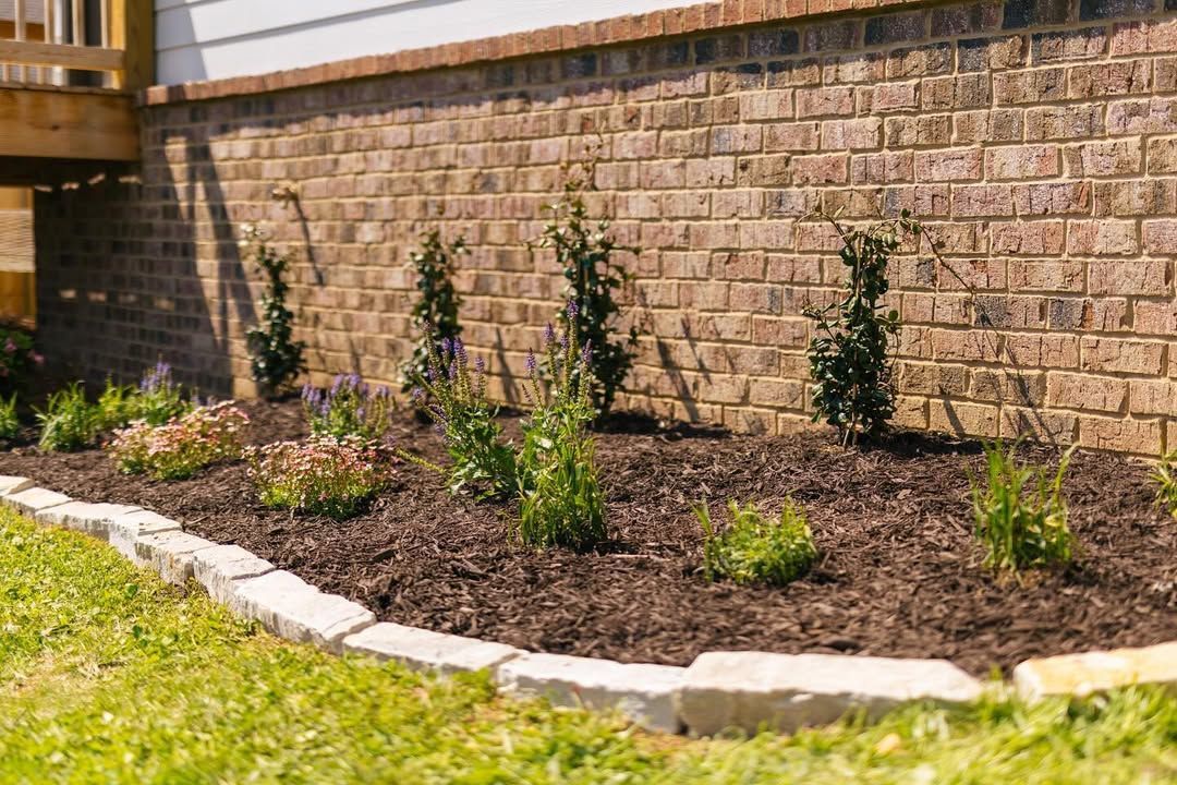 Brick foundation with landscaped bed of plants and mulch, bordered by stone edging.