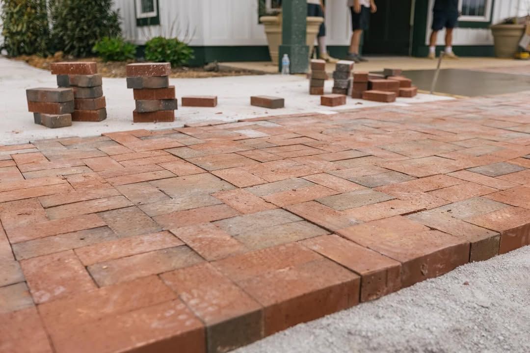 Red bricks being laid in a herringbone pattern on a sandy surface, near a building.