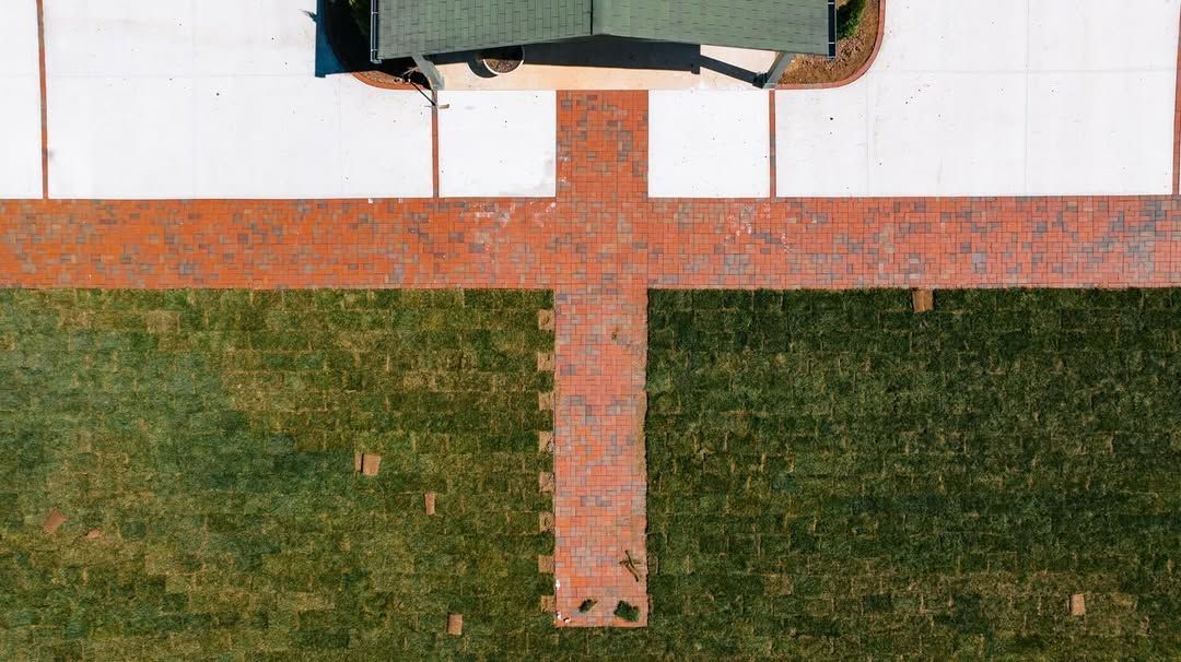 Overhead view of a grassy lawn with a red brick pathway leading to a driveway and a portion of a house.