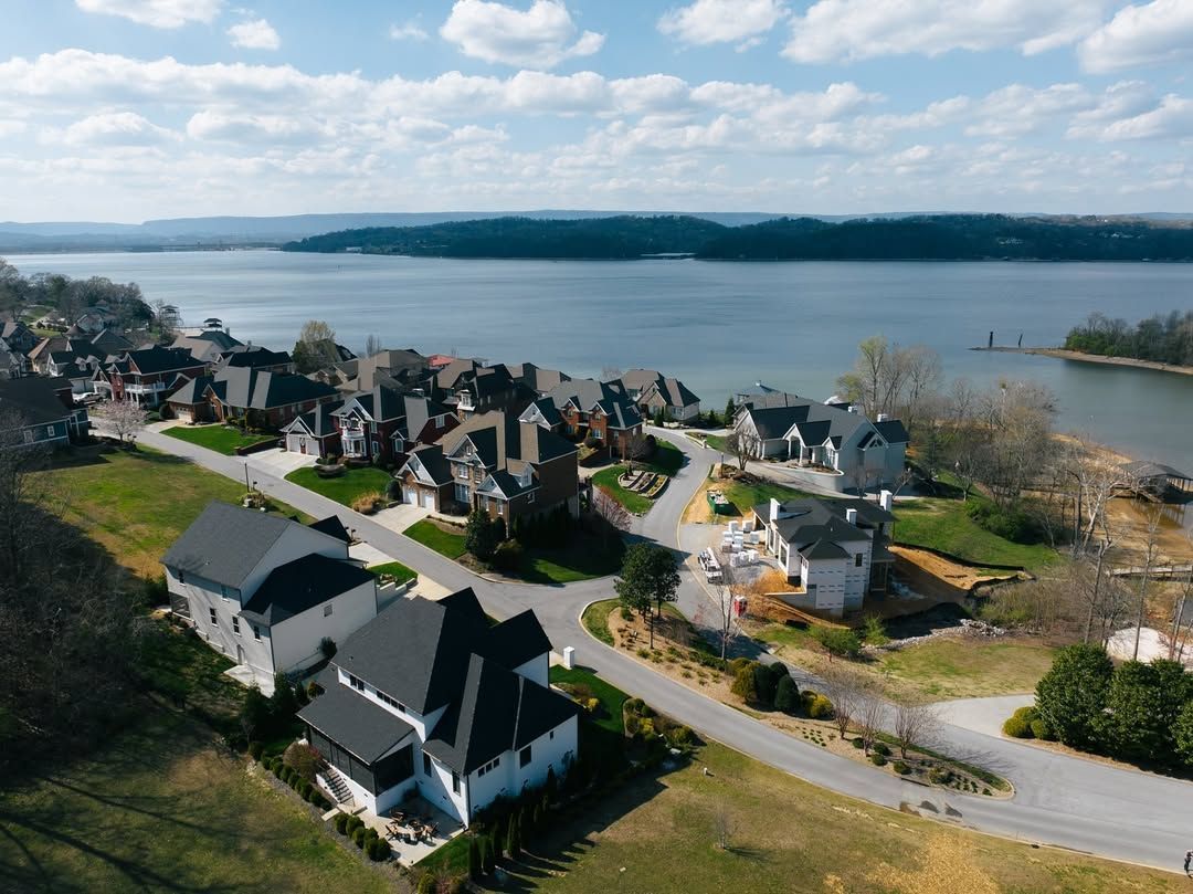 Aerial view of houses along a waterfront road, with a large lake and forested land in the background.