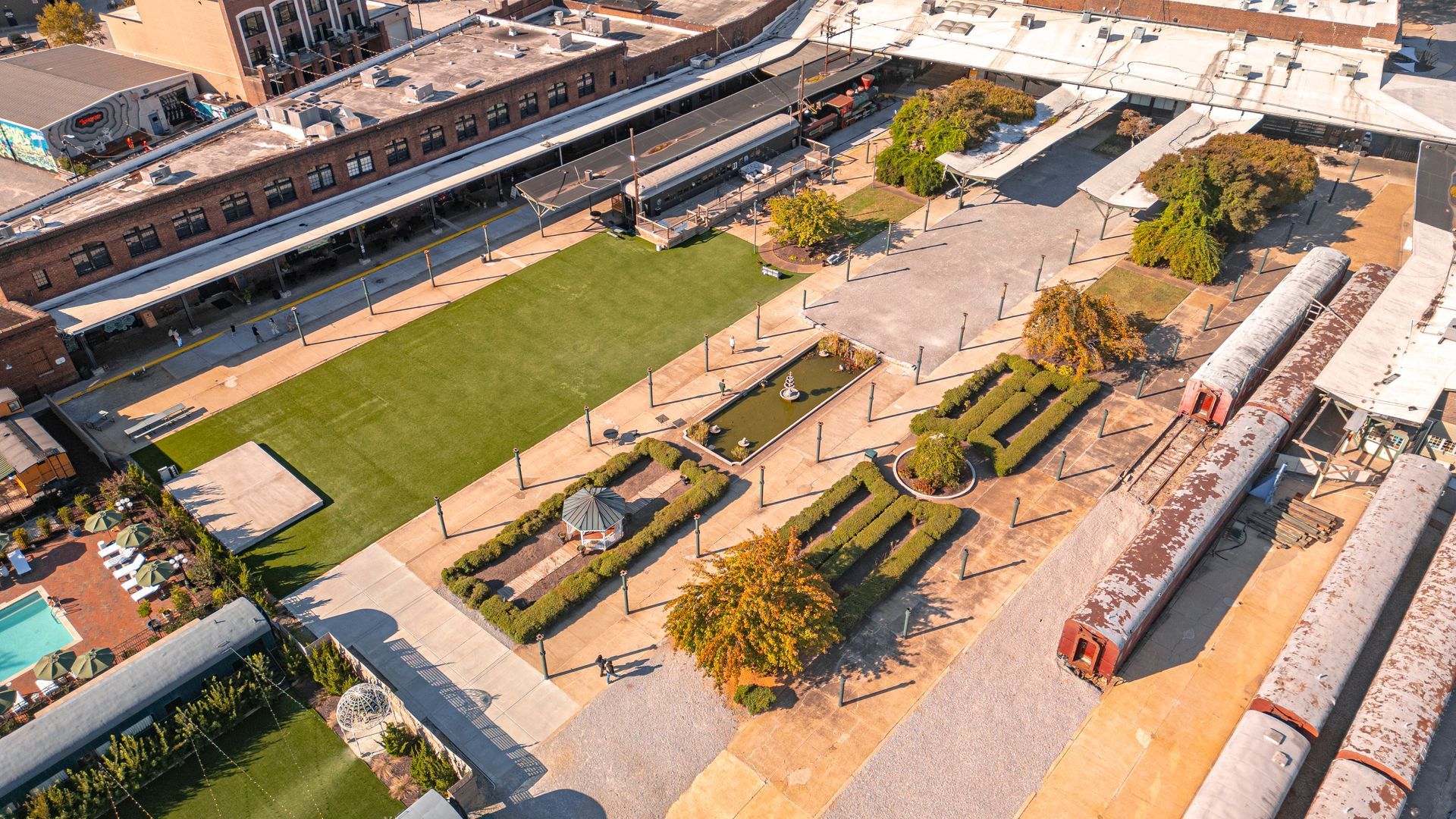 Aerial view of a plaza with landscaping, a grassy area, and train cars, adjacent to brick buildings.