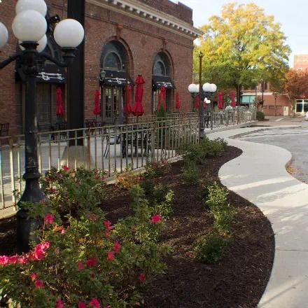 A brick building with red umbrellas and a patio with a curved sidewalk lined with bushes and lamp posts.
