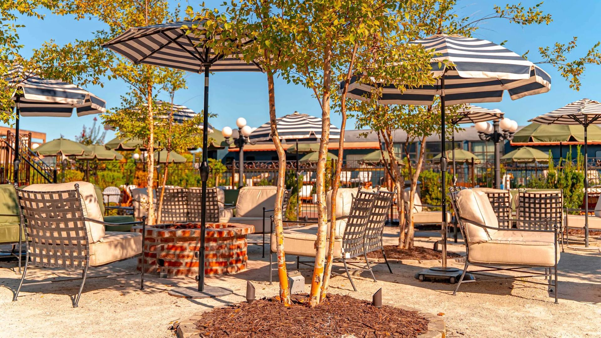 Outdoor patio with striped umbrellas, lounge chairs, a fire pit, and small trees.