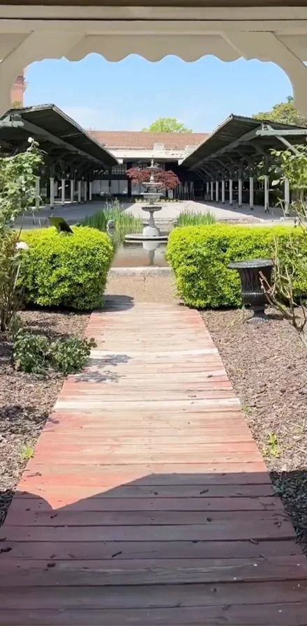 A wooden walkway leads to a garden with green hedges, a fountain, and covered structures under a blue sky.