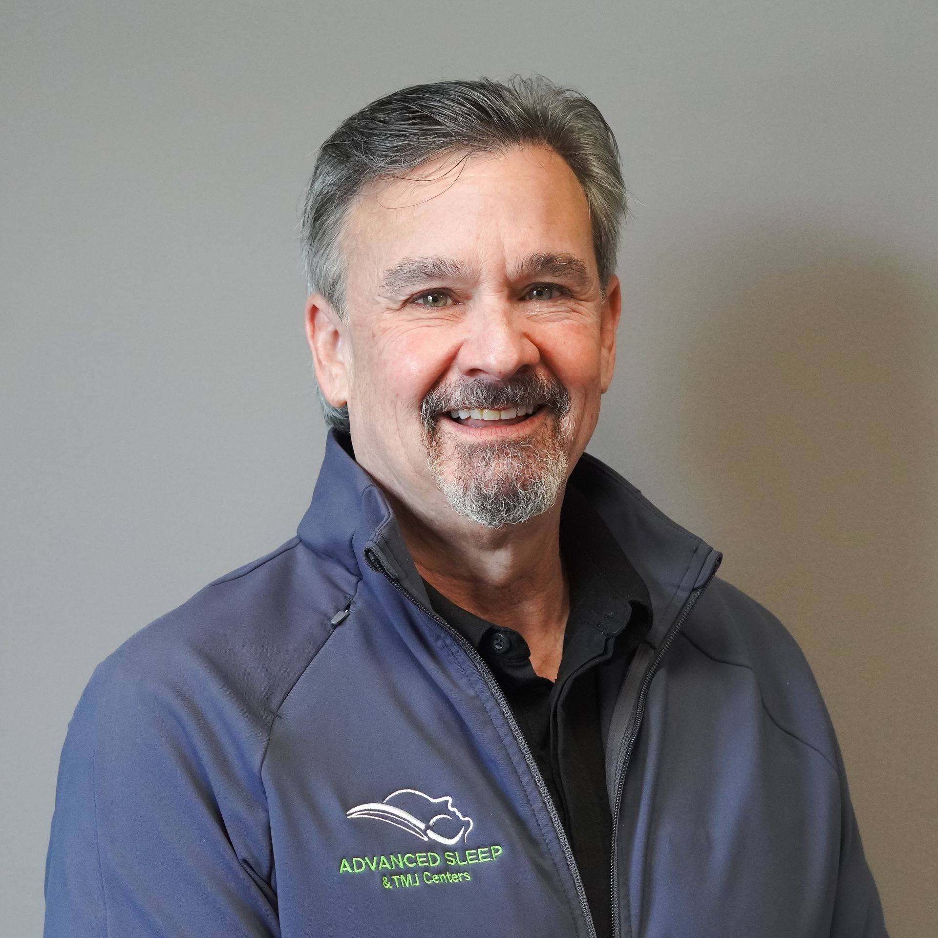 A man with a beard is wearing a blue jacket and smiling.