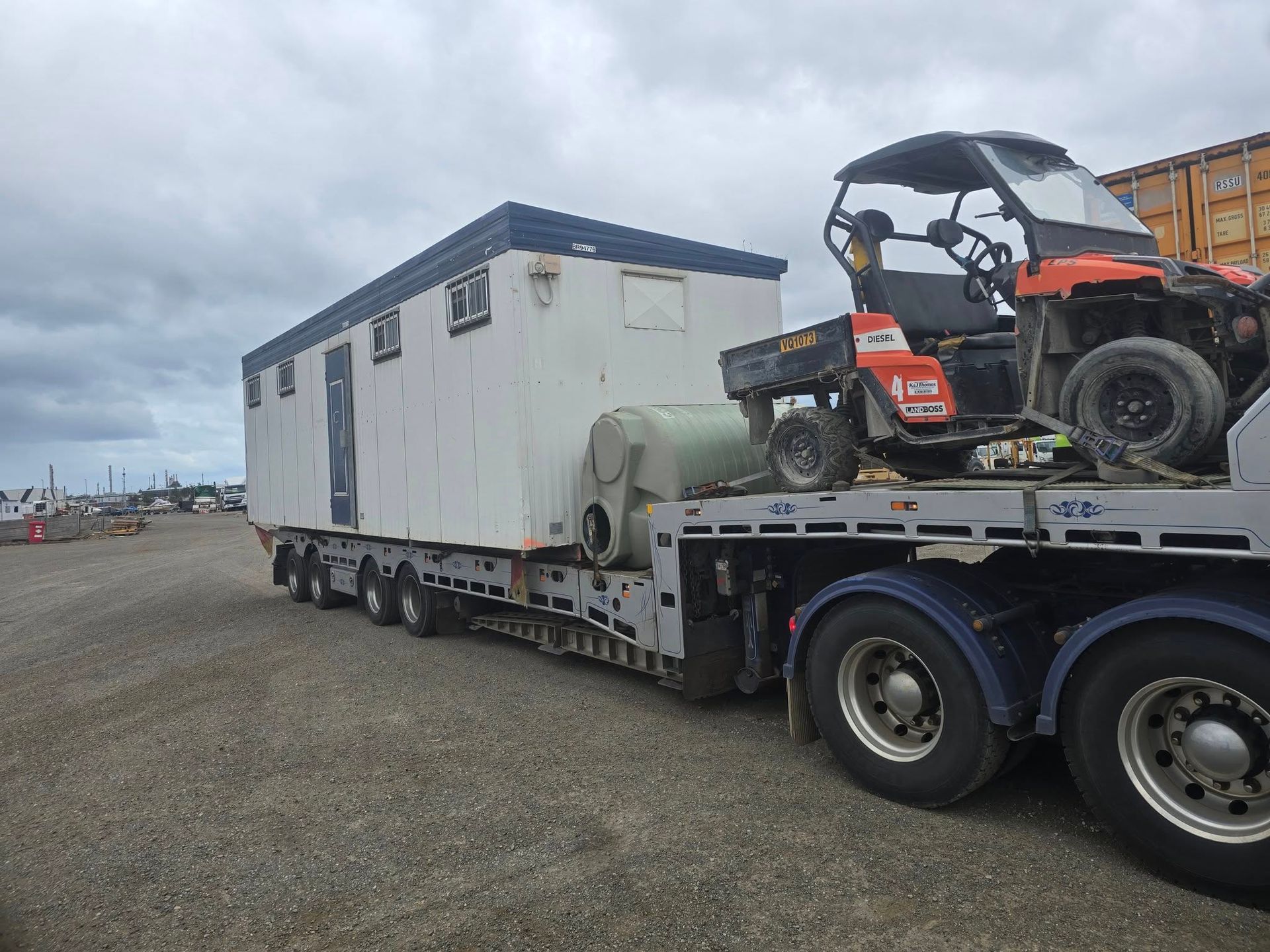 Flatbed truck hauling a white portable building and an orange ATV on a gravel lot under cloudy skies  — Ballarat Supatilt in Delacombe, VIC