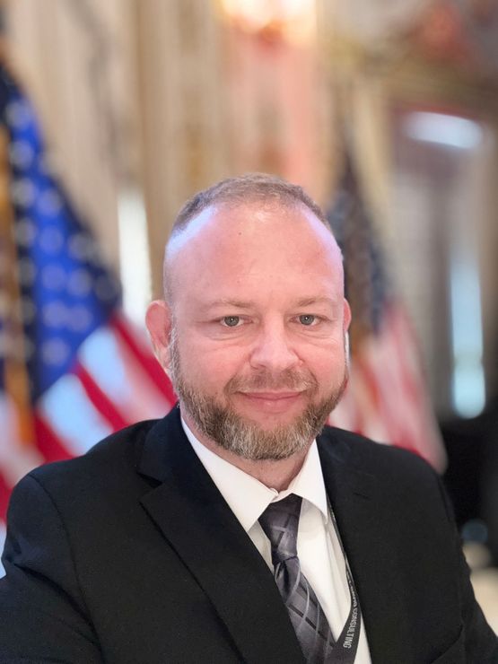 A man in a suit smiles at the camera in front of American flags, likely in a government building.