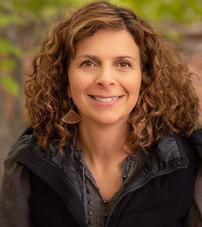 A woman with curly hair is wearing a black vest and smiling for the camera.