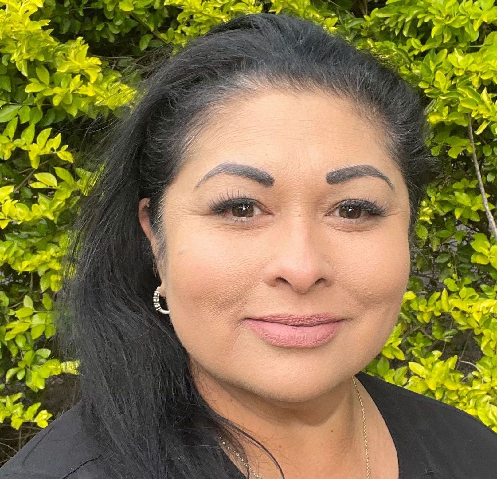 A woman with long black hair is smiling for the camera in front of a bush.