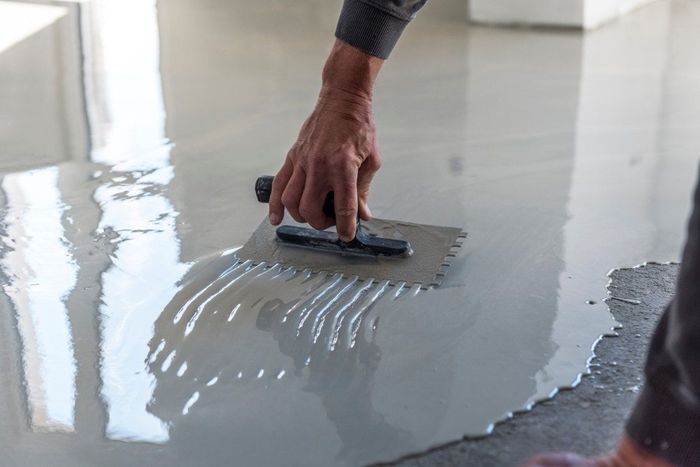 Person using a notched trowel to spread gray mortar on a concrete floor.