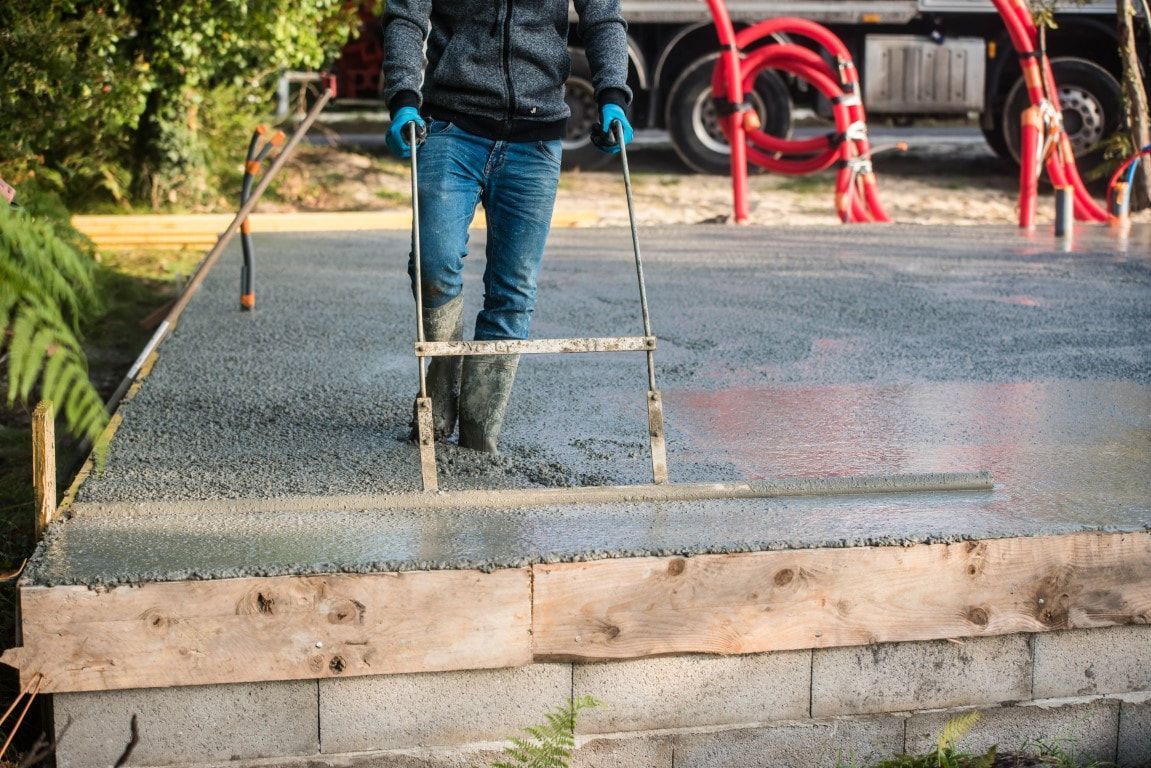 Man leveling wet concrete on a foundation. Construction site with truck and red hoses visible.