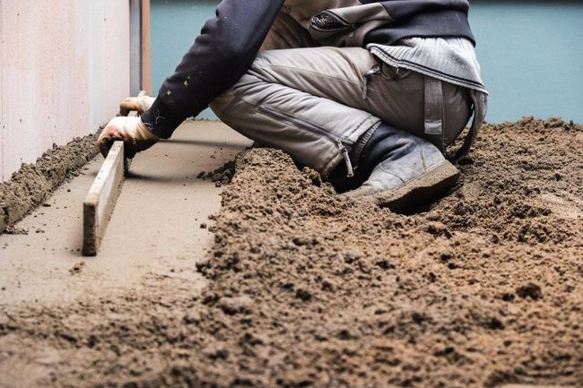 Person kneeling, leveling wet cement with a board. Muddy ground, light brown cement, indoors.