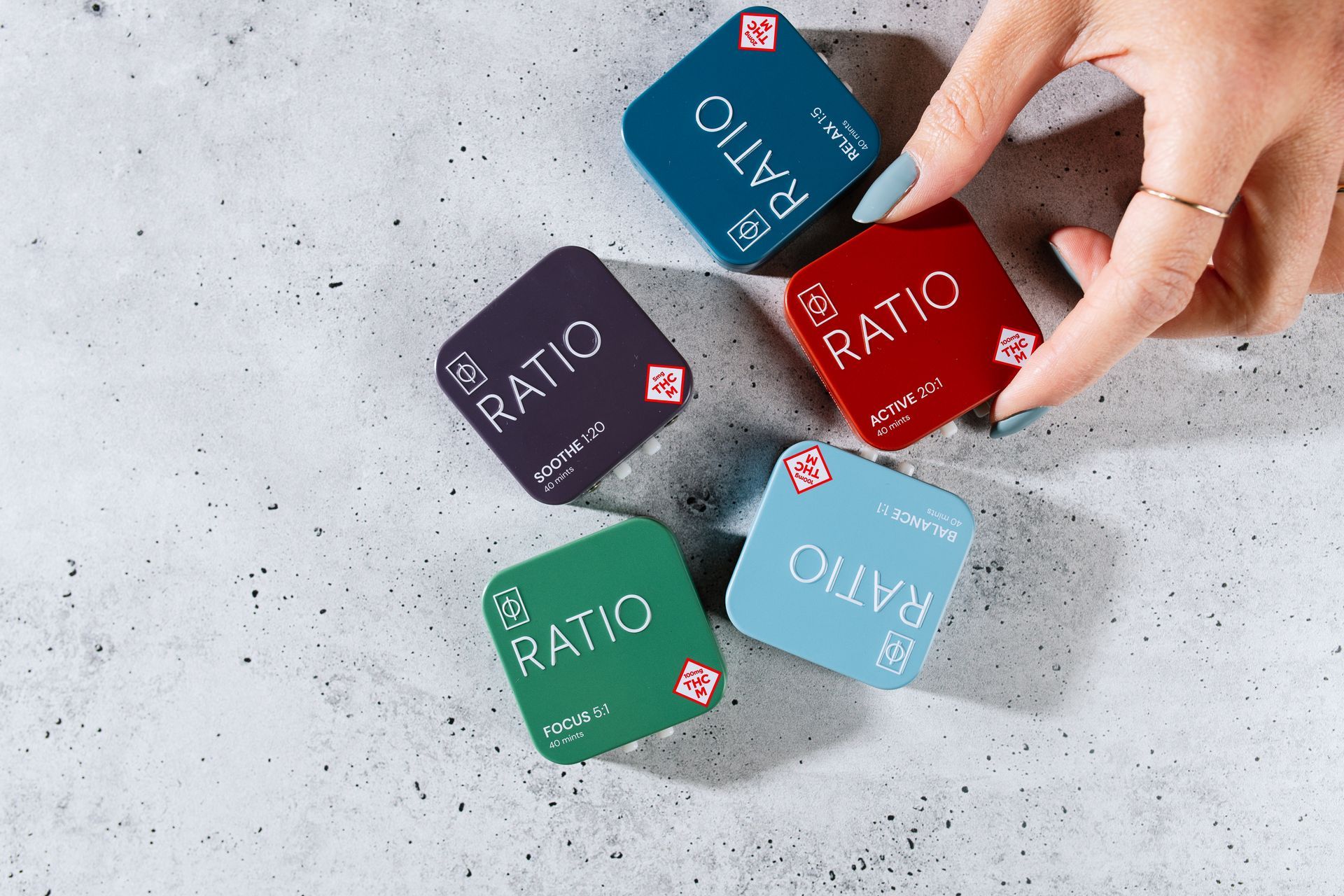 Red, blue, purple, green, and light blue RATIO tins on cement background with hand reaching for it.
