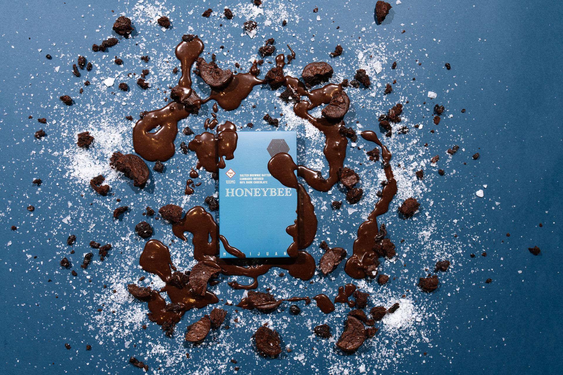 Salted Brownie Batter packaging on a blue background with sea salt and brownie bits with brownie batter drizzled on top.