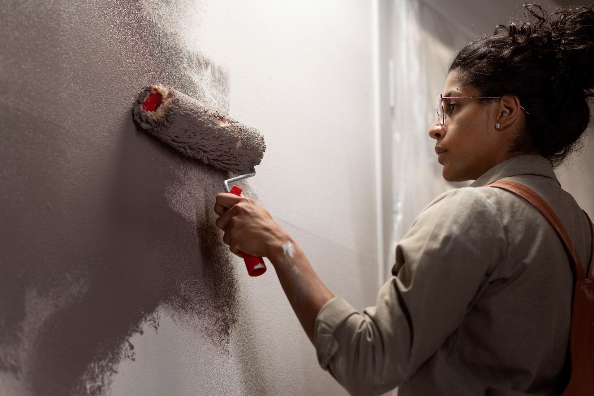 Person paints a wall gray with a roller. Interior setting, visible shoulder bag.