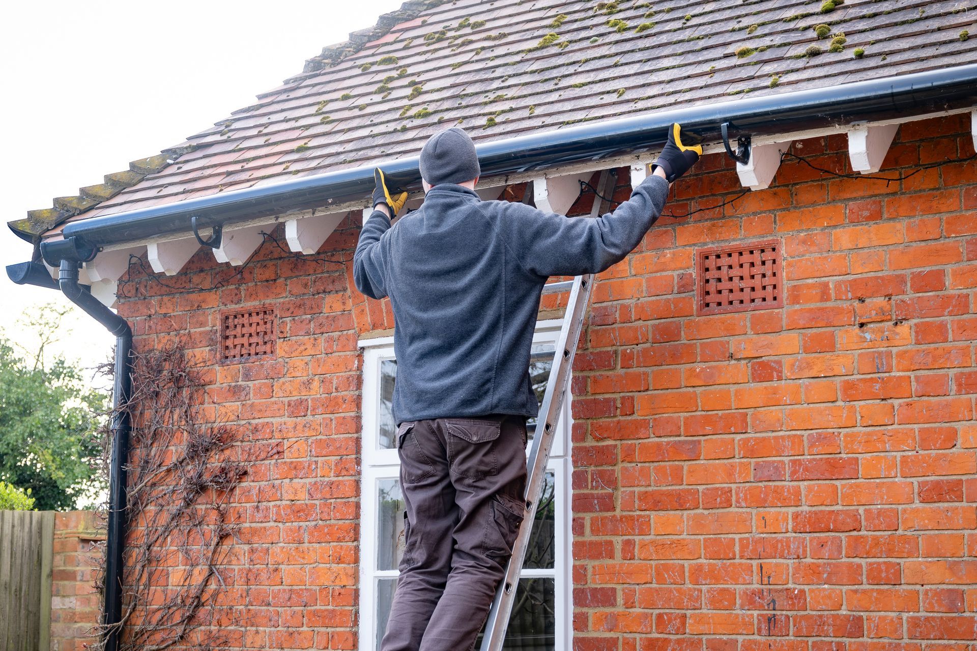 Man on a ladder cleaning gutters of a brick house with a tiled roof.