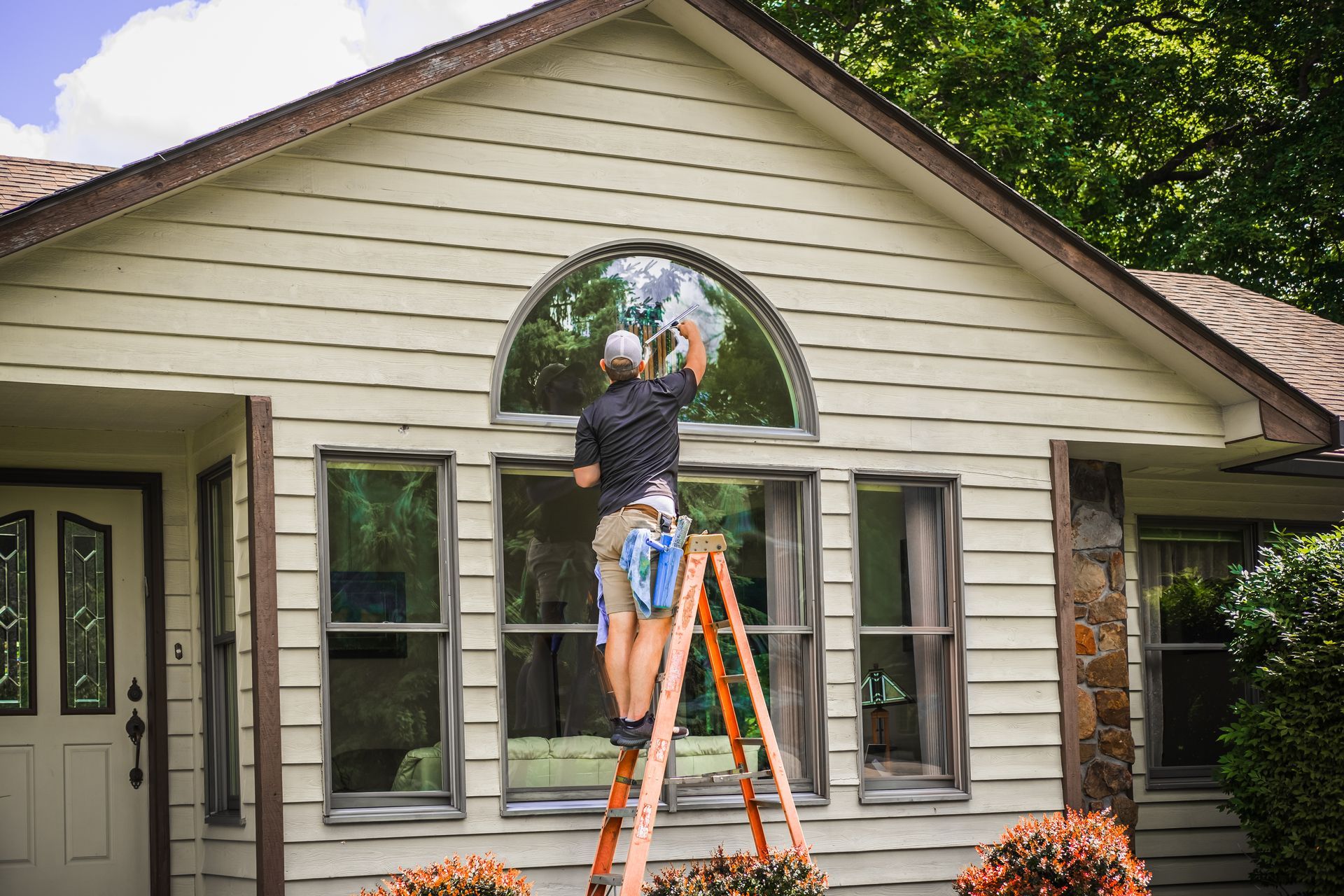 A man on a ladder is cleaning the windows of a brick house.