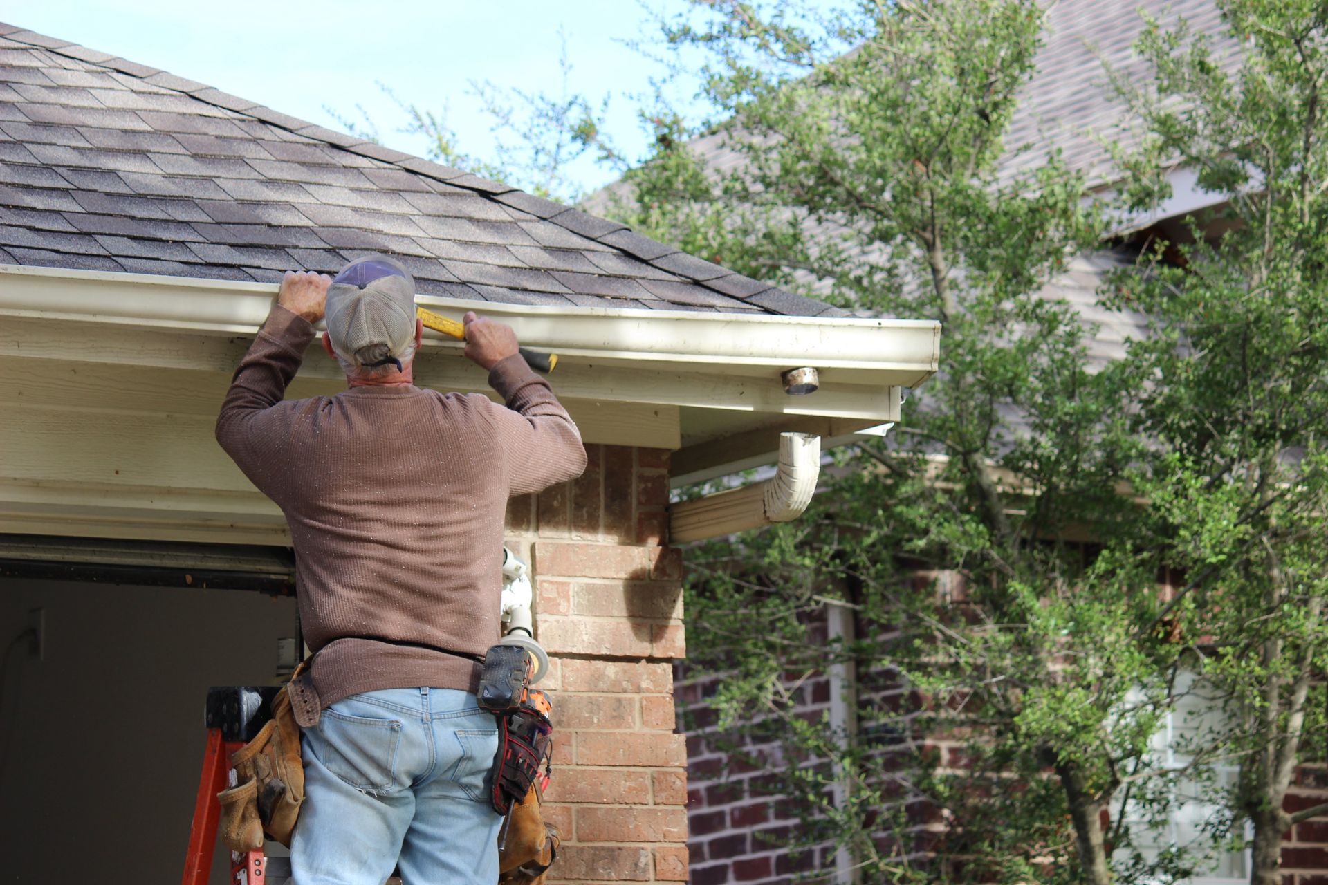 A man is standing on a ladder fixing a gutter on the roof of a house.