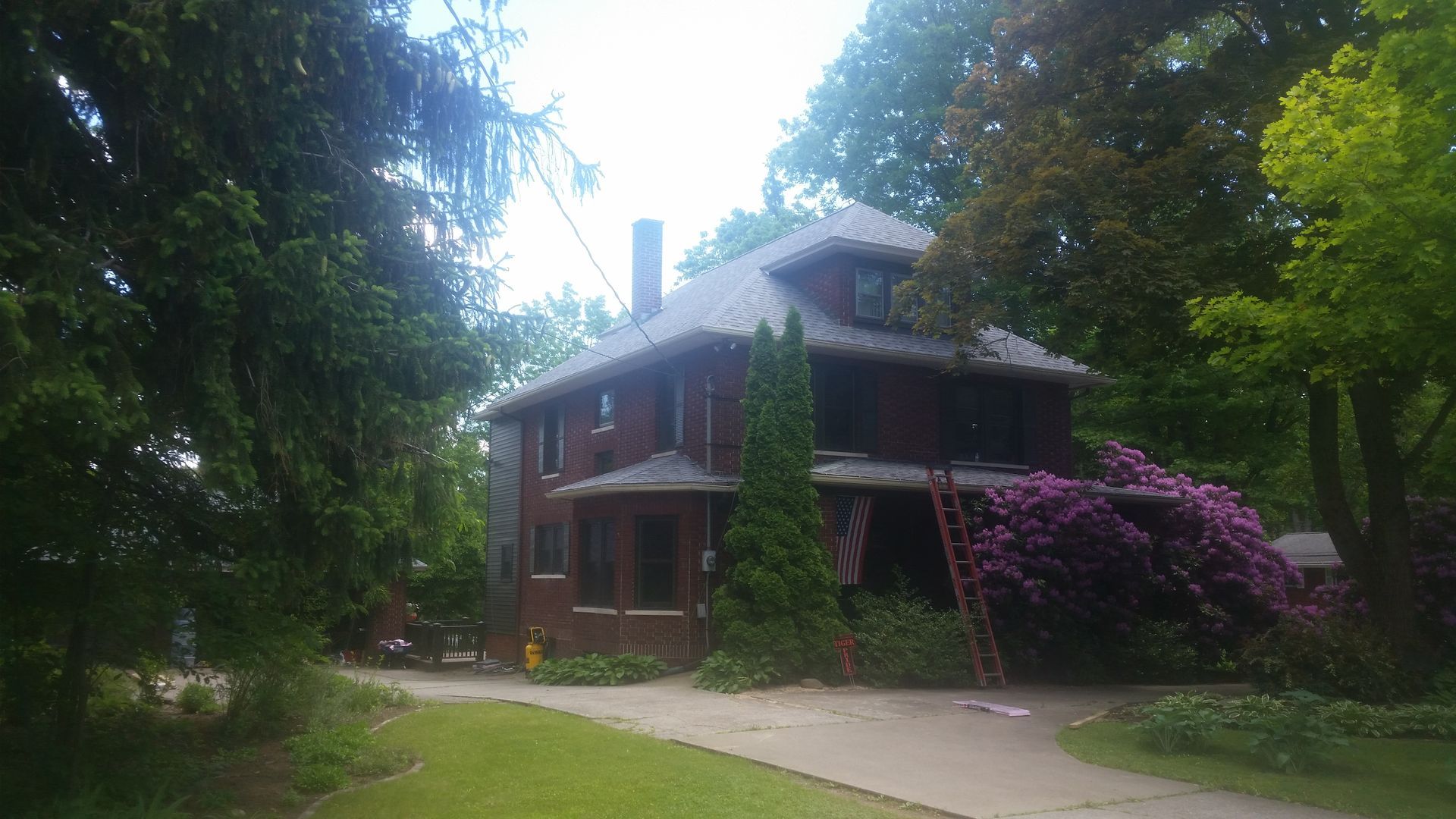 A large brick house with a ladder in front of it is surrounded by trees.
