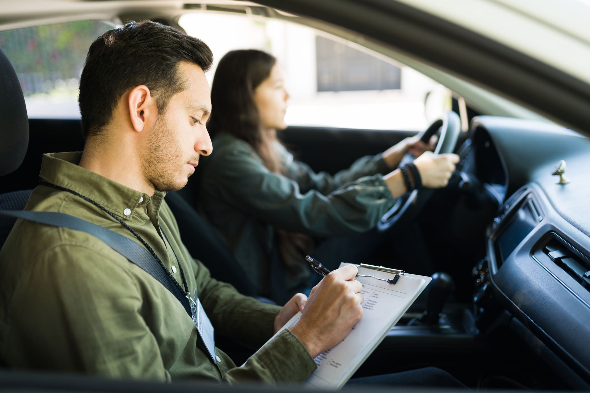Driving instructor in passenger seat, taking notes as driver steers vehicle.