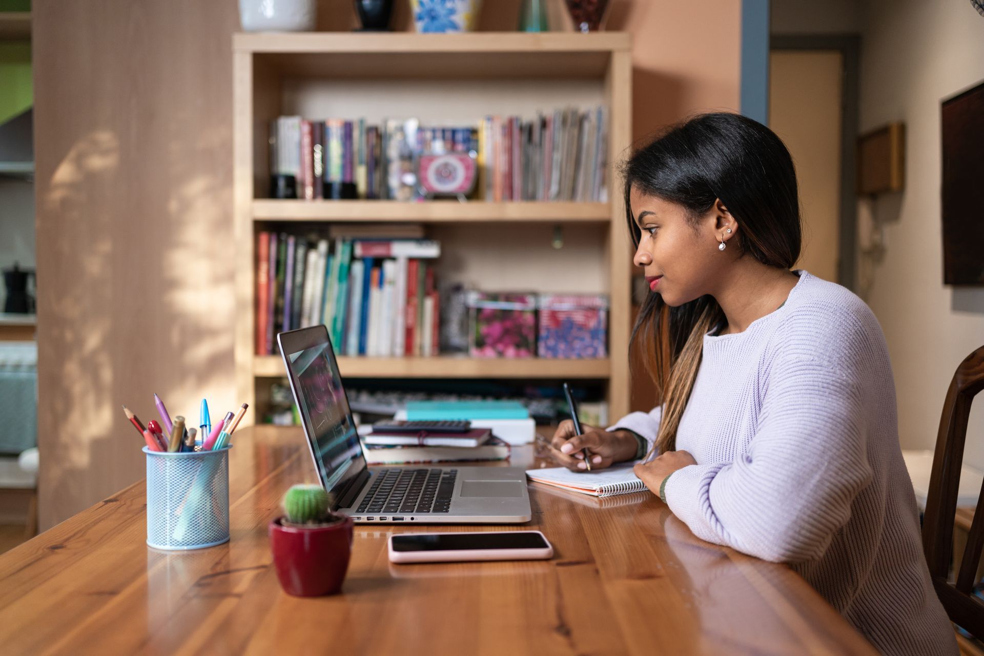 Woman working on laptop, taking notes at a wooden table in a home office.