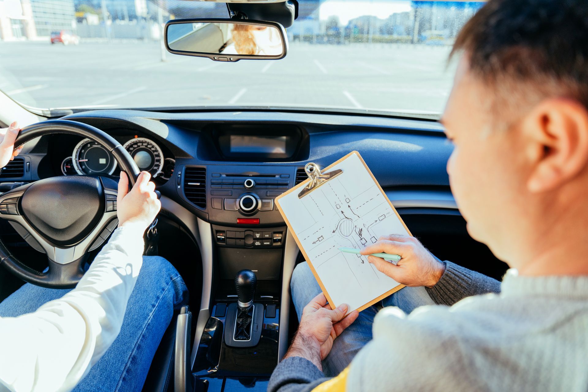 Driving lesson: instructor points at diagram on clipboard, student holds steering wheel. 