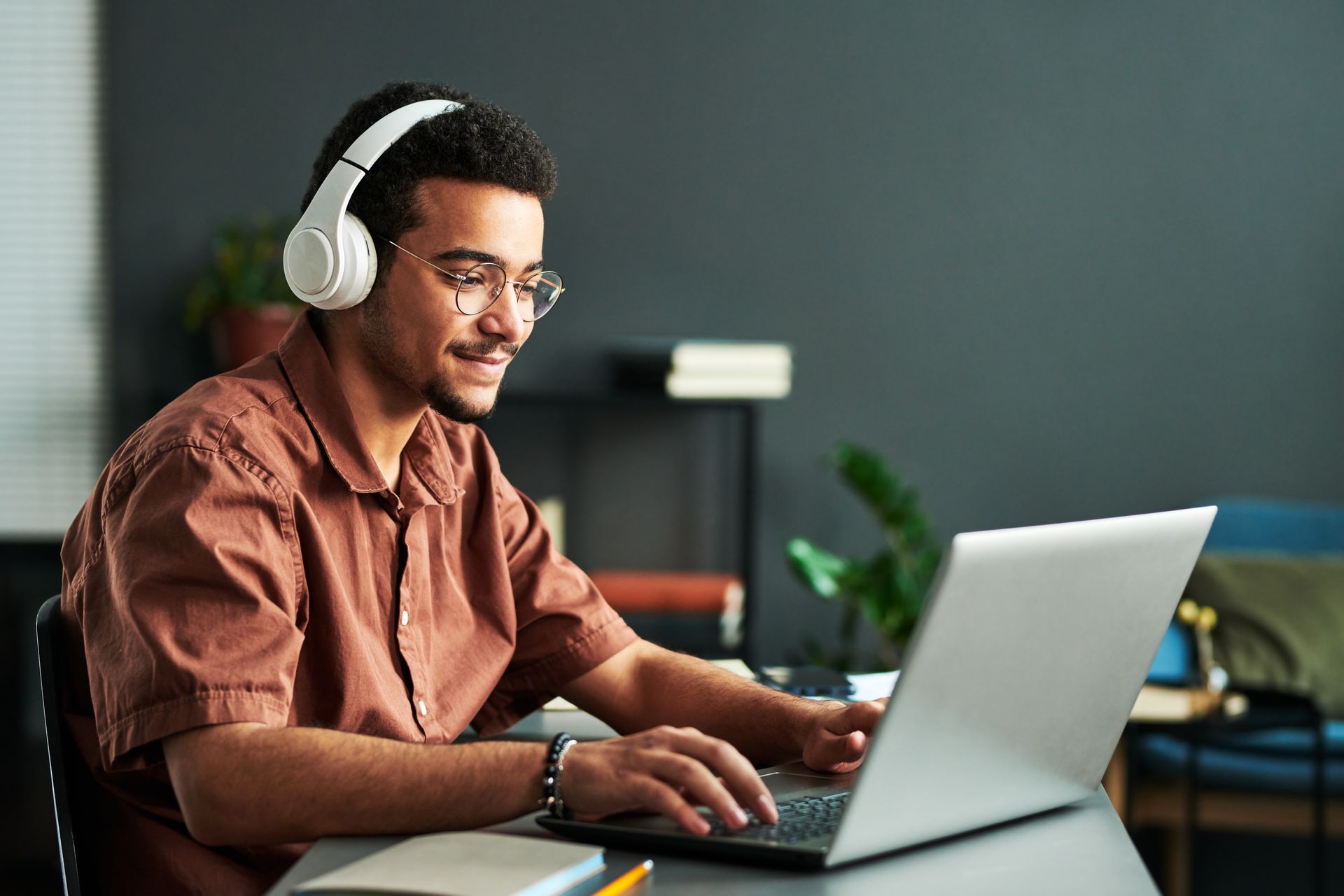 Man wearing headphones and glasses, working on a laptop at a desk, smiling.
