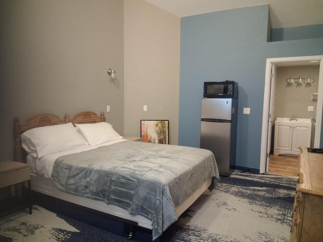 Bedroom with bed, refrigerator, and bathroom doorway. Blue and gray walls and carpet.