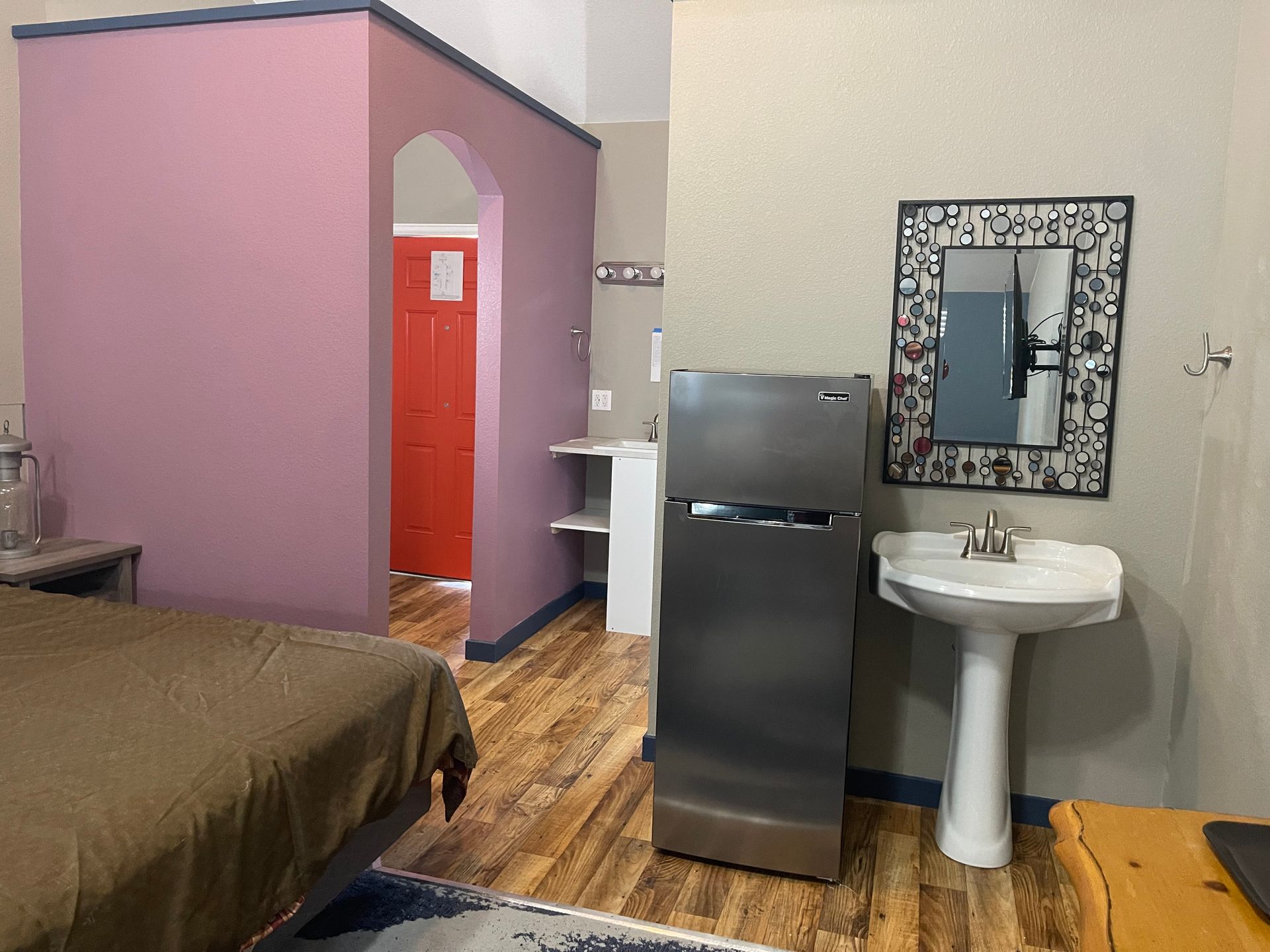Bedroom with bed, mini-fridge, sink, mirror, and pink archway leading to a red door.