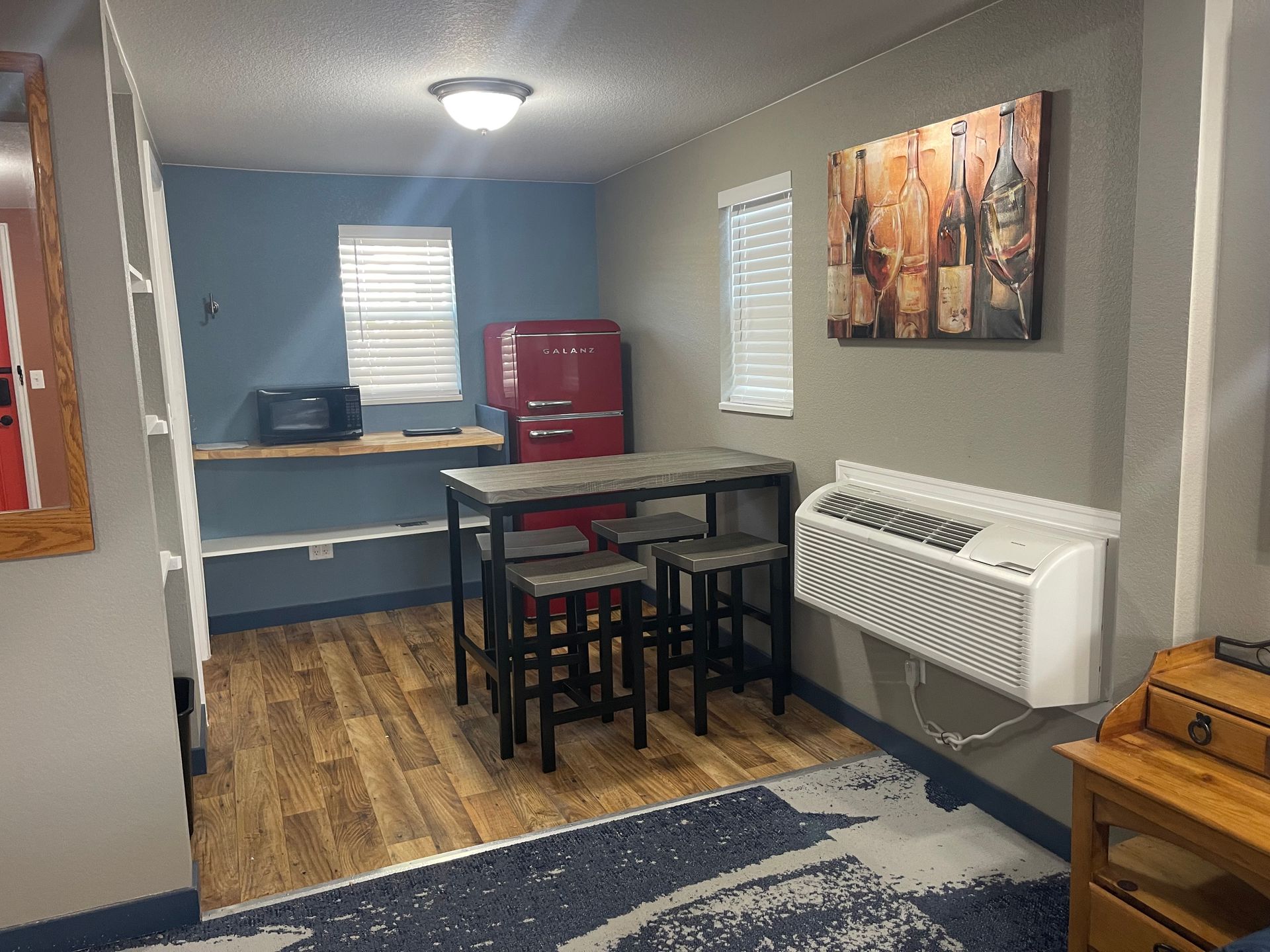 Small studio kitchen: dining table with stools, mini fridge, microwave, shelves, and blue wall.