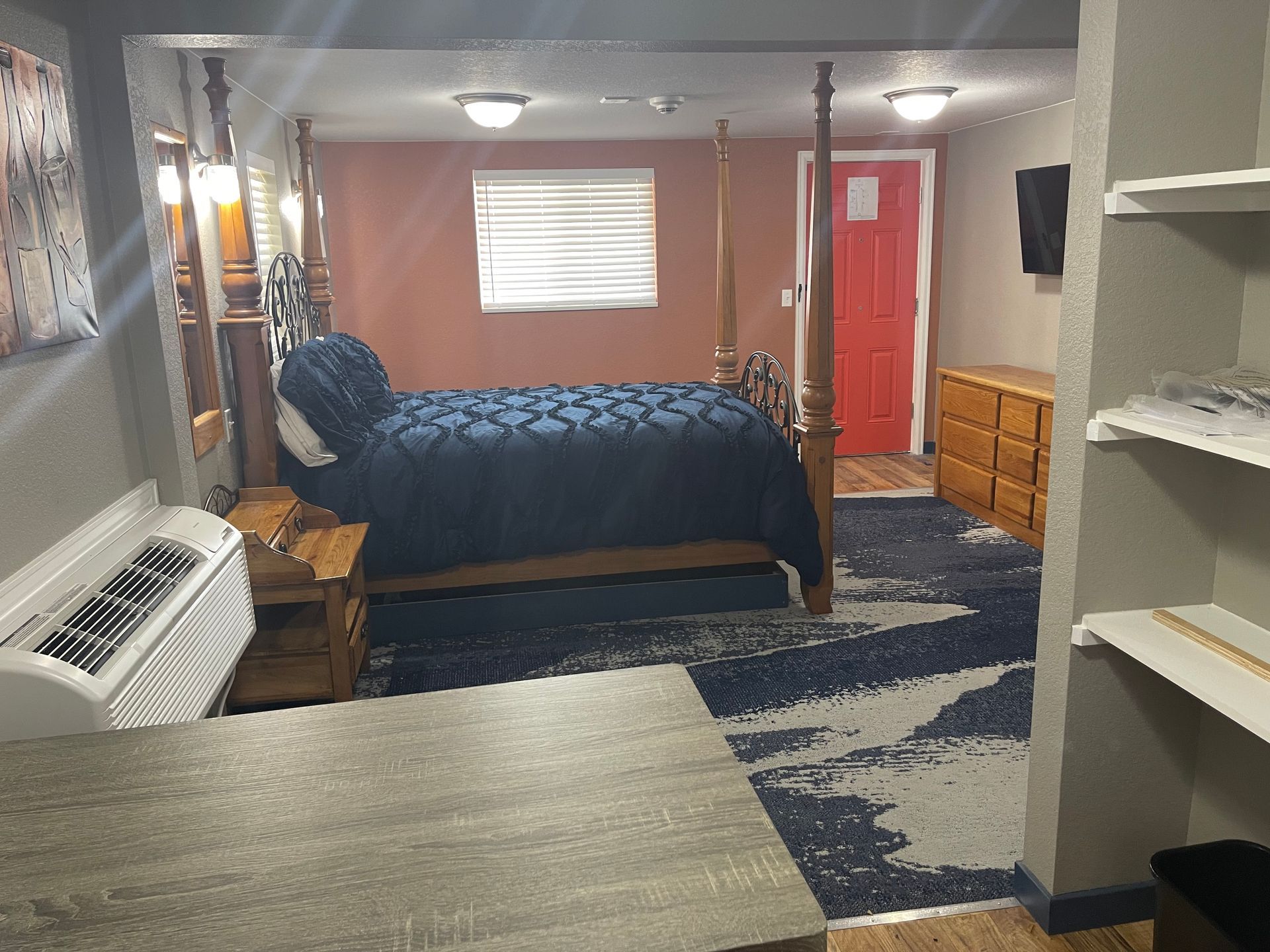 Hotel room with a bed, desk, and a large window. Wood paneling and neutral colors.