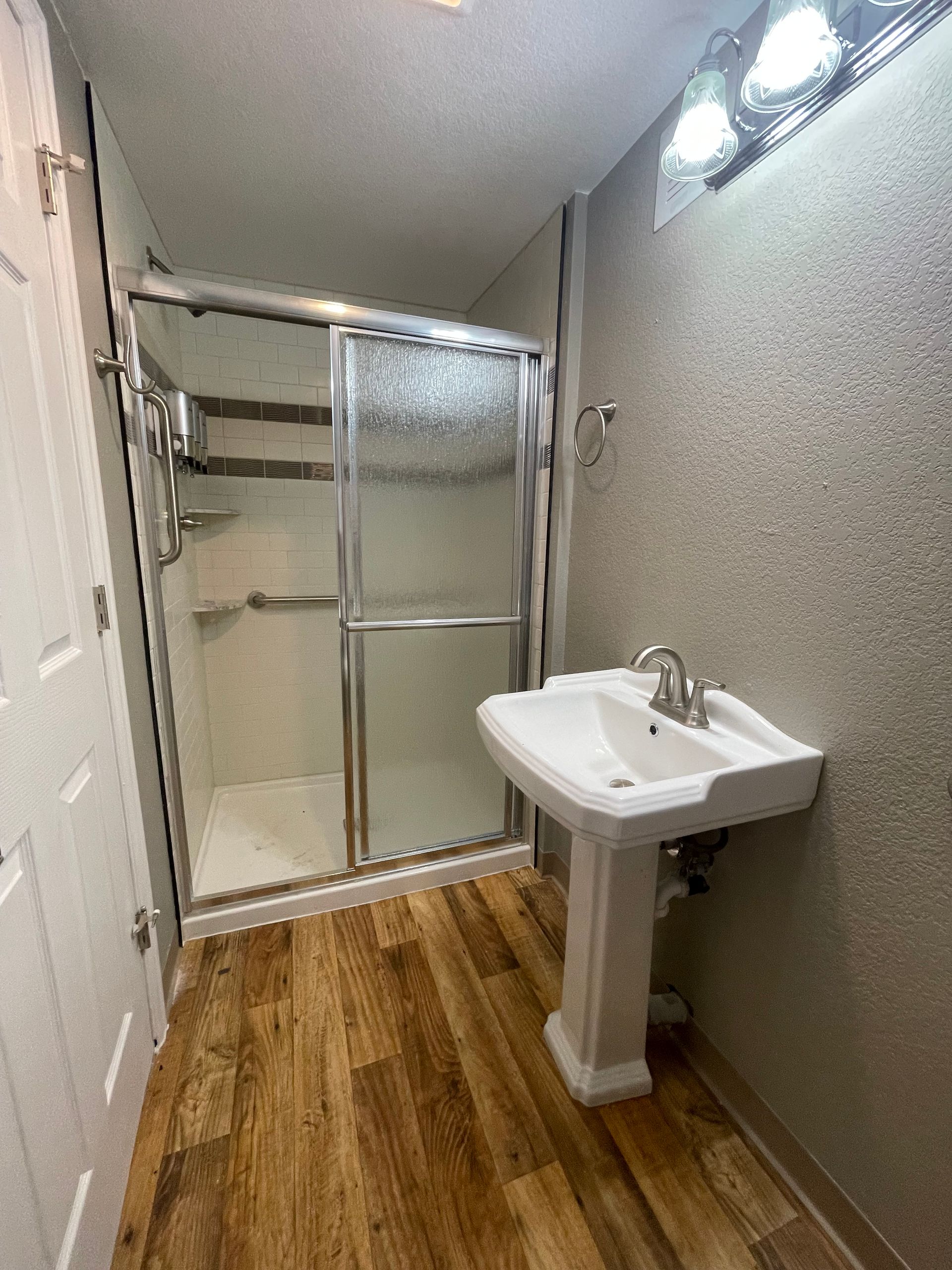 Bathroom with shower, pedestal sink, and wood-look flooring. Shower has glass doors. Light fixture above sink.