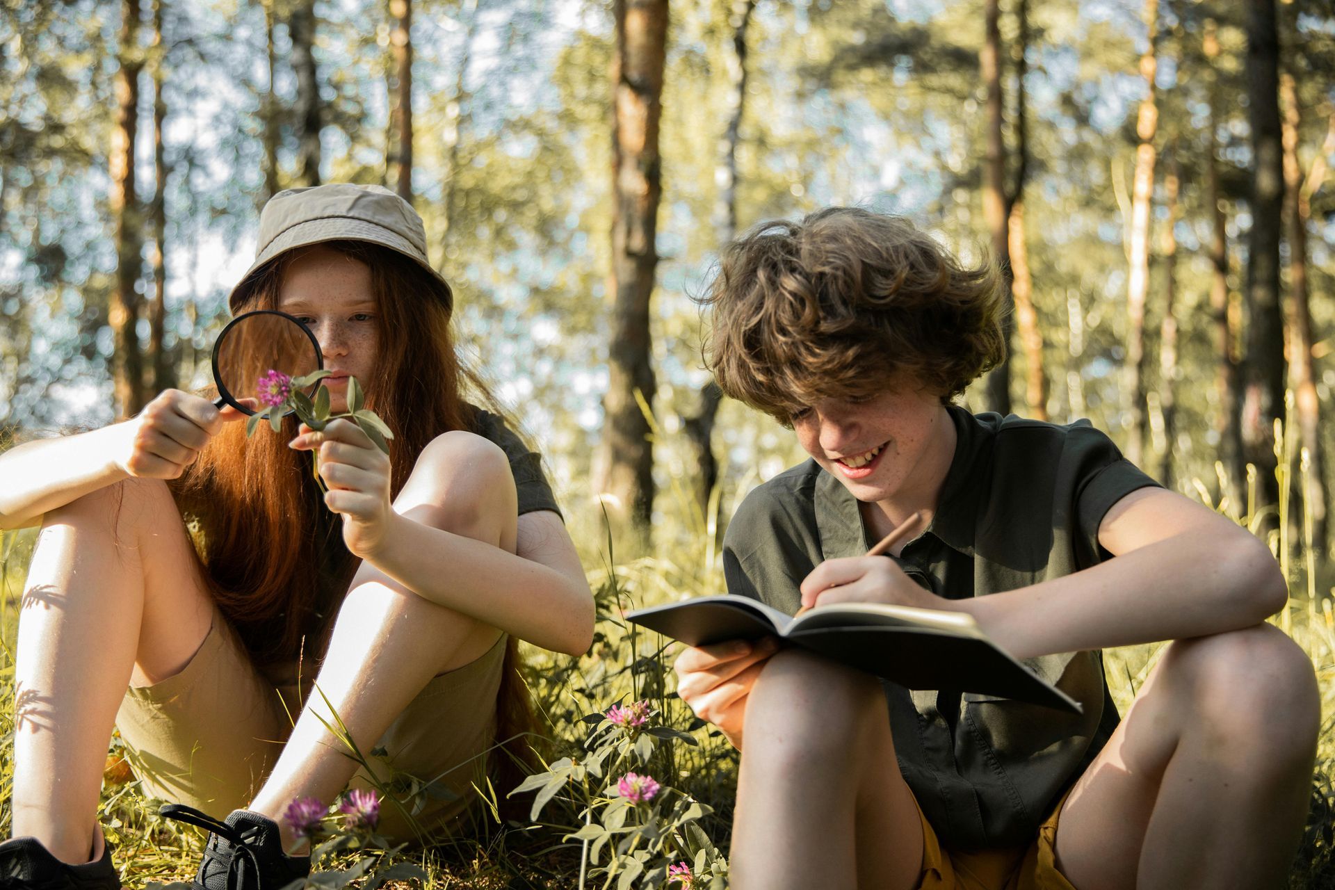 Two kids in a forest, one using a magnifying glass on a flower, the other writing in a notebook.