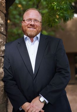 A man in a suit and glasses is standing next to a tree.