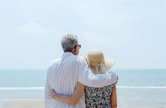 An elderly couple is standing on the beach looking at the ocean.