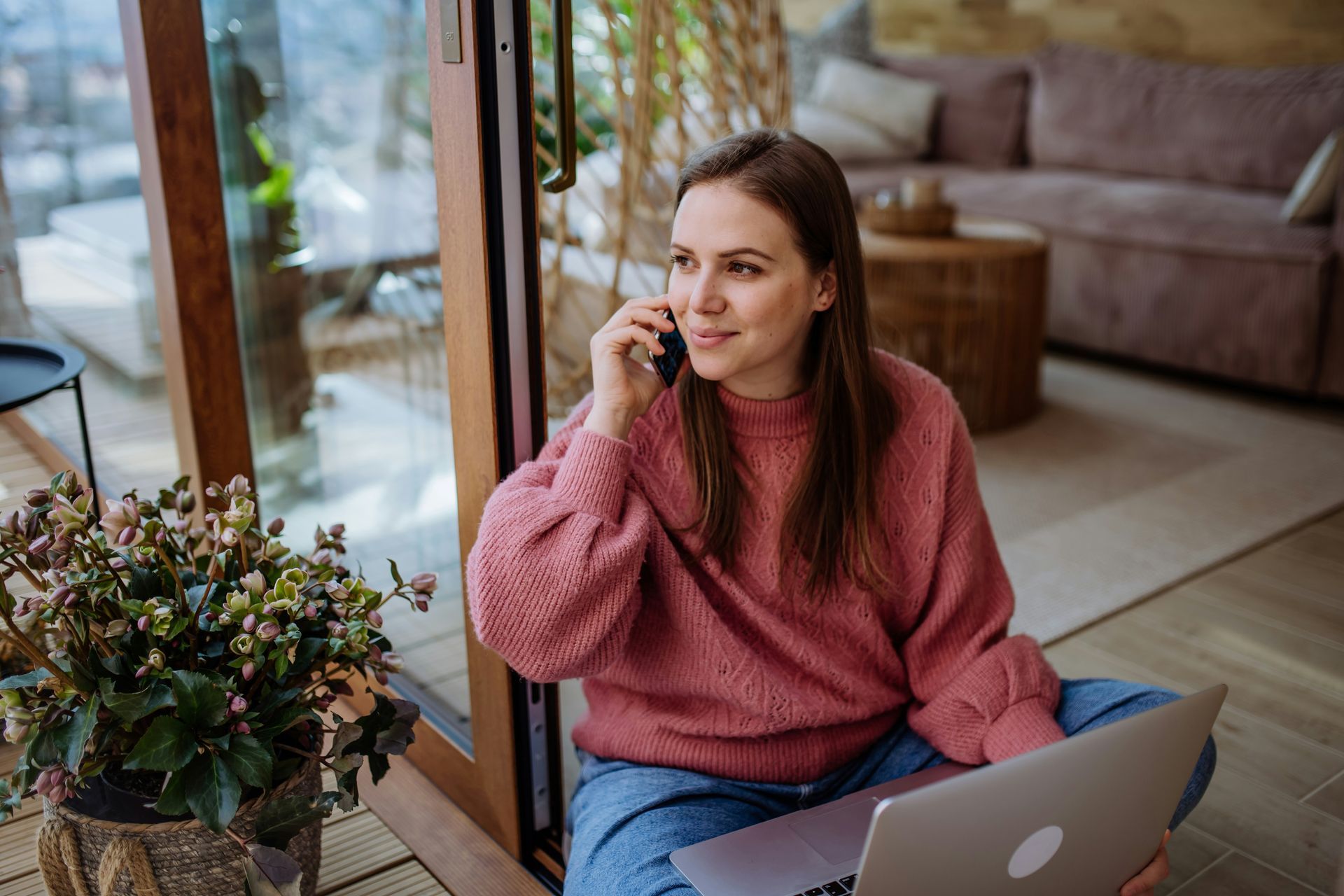 A woman is sitting on the floor with a laptop and talking on a cell phone.