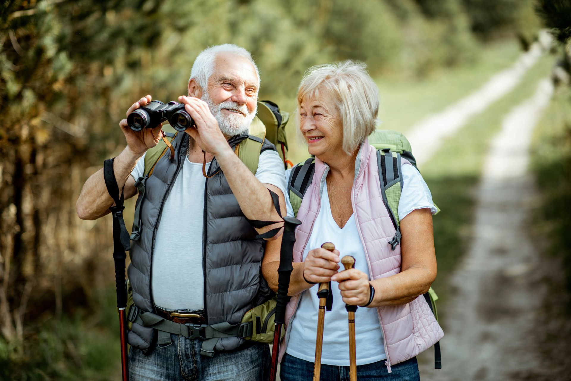 An elderly couple is hiking in the woods and looking through binoculars.