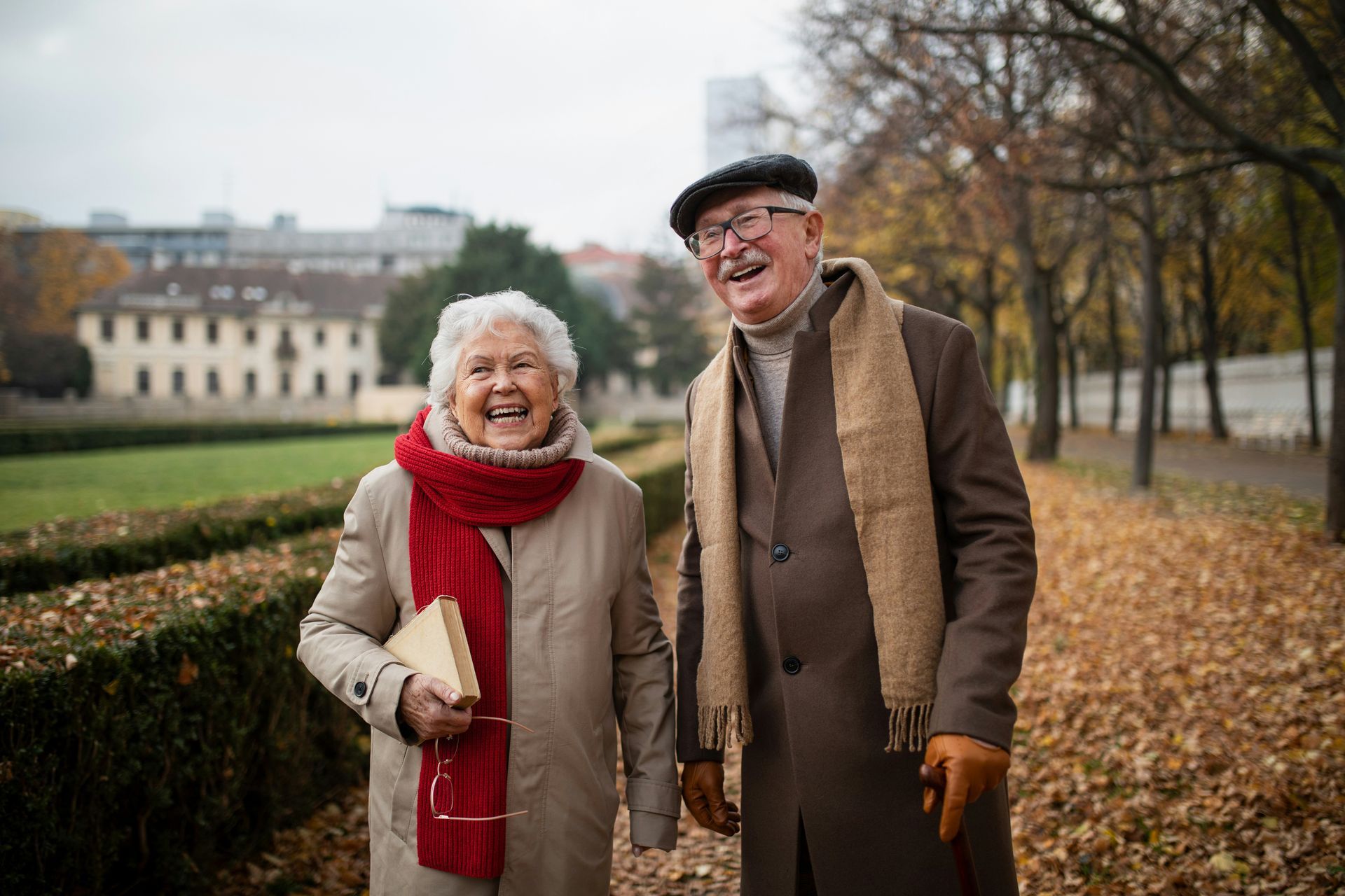 An elderly couple is walking in a park holding hands and smiling.