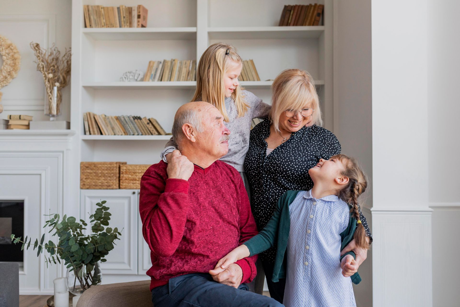 Four people smiling in a bright living room with shelves and a fireplace; one child stands between adults and another leans back.