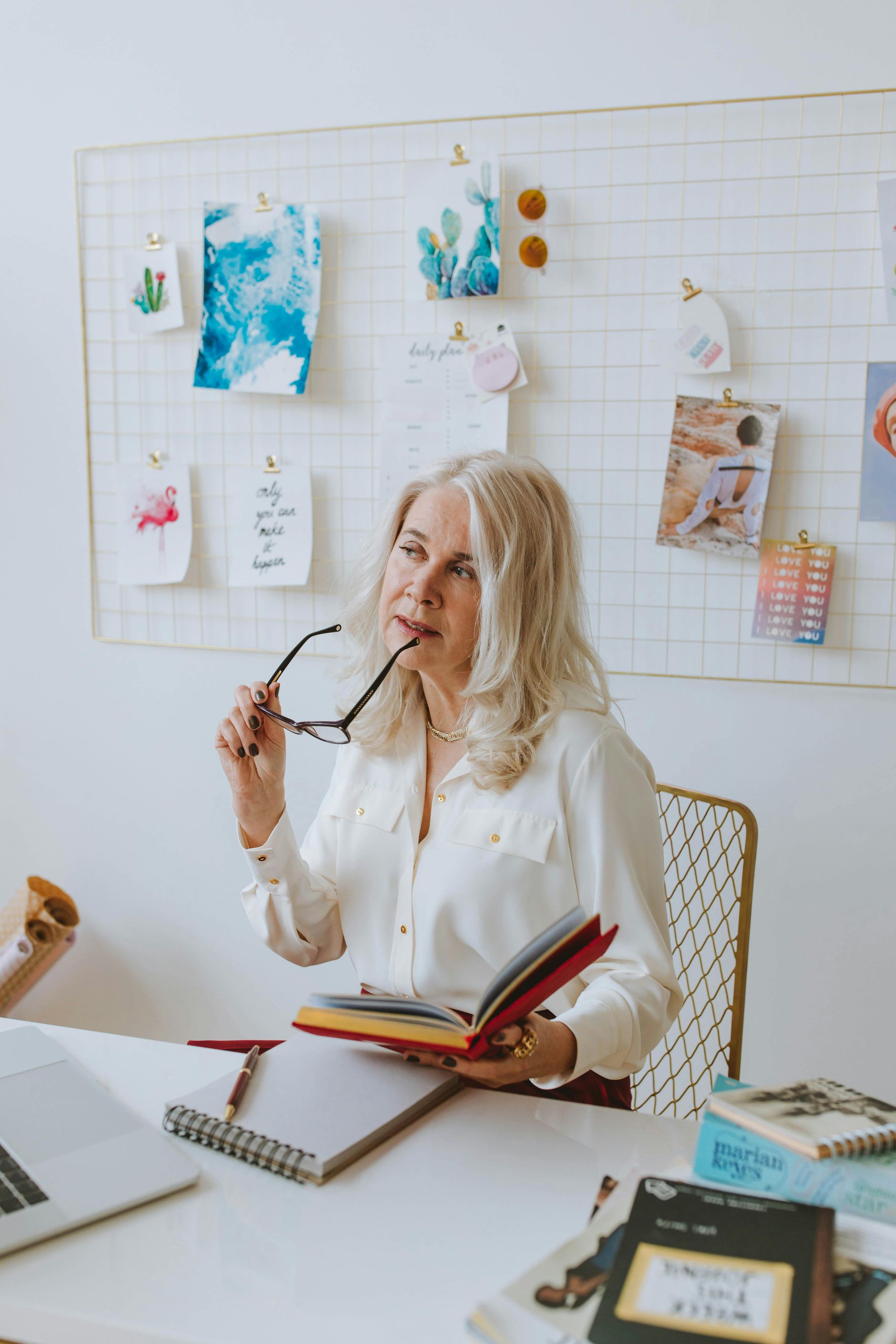 A woman is sitting at a desk wearing glasses and holding a book.