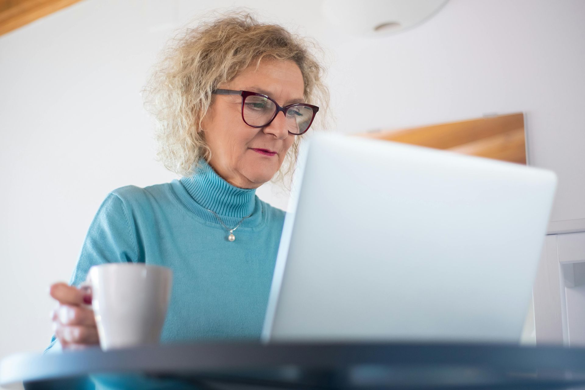 A woman is sitting at a table using a laptop computer while holding a cup of coffee.